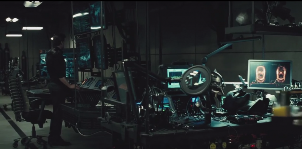 Batman aka Bruce Wayne next to the chair and desking in the Batcave